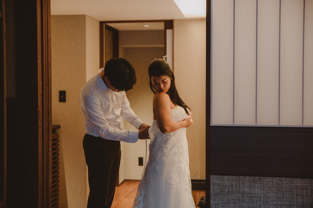 Japan Roadtrip Elopement- The groom is zipper up the bride's wedding dress 