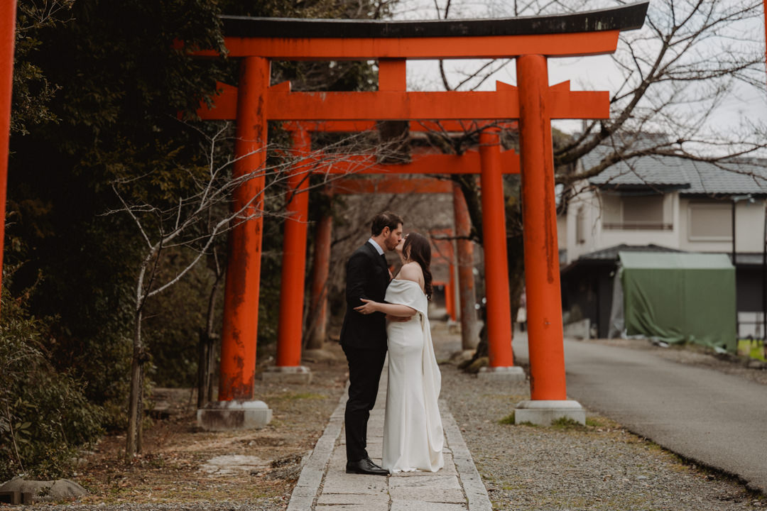 The couple kisses under a red torii gate. Snow Shrine Elopement in Japan